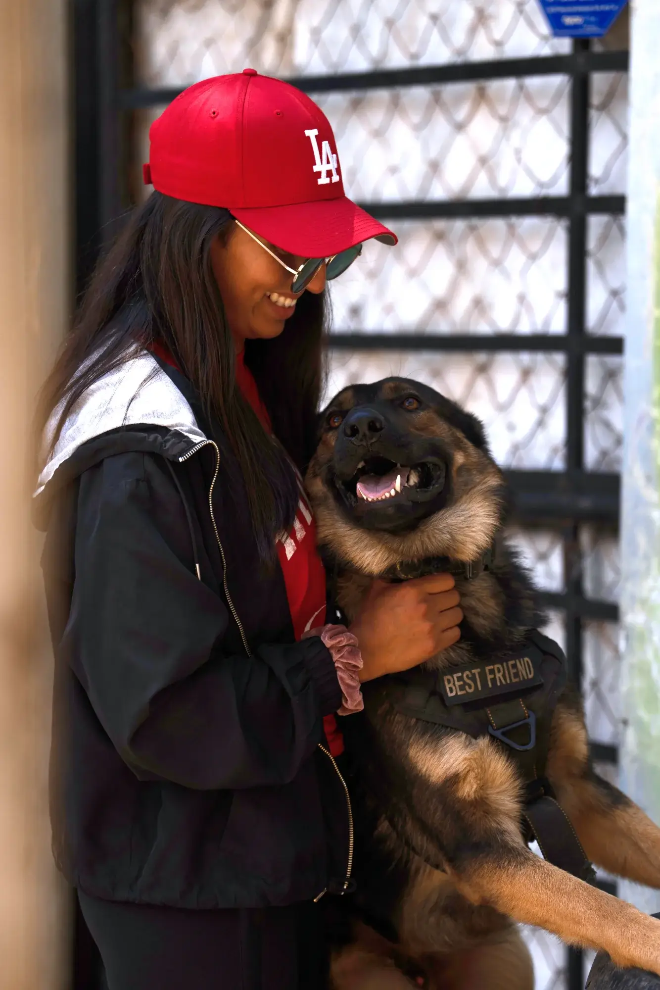 Una persona con gorra roja y gafas de sol sonríe mientras abraza a un perro grande con un arnés que dice Mejor Amigo. Están de pie cerca de una puerta, y el perro parece feliz y contento.